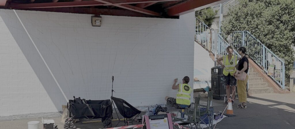 Katie and Iona talking at the base of the bridge while Matt from Art Streets works on the outline of the mural